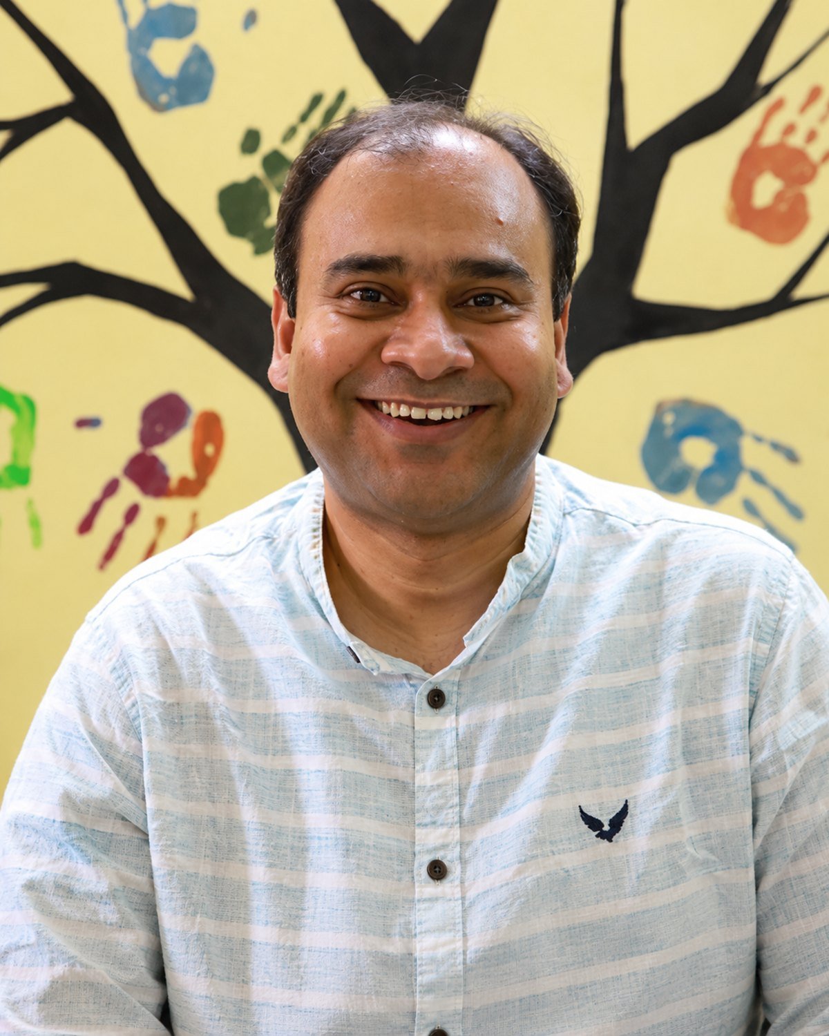 Bappaditya Mukherjee, photographed in front of a painted tree mural whose branches are made of children's coloured handprints.